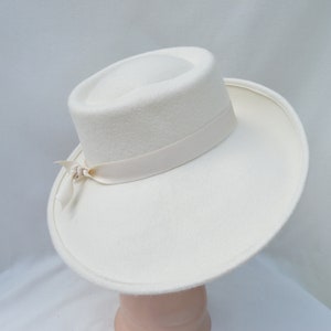 May include: A cream-colored hat with a wide brim and a cylindrical crown. A cream-colored ribbon is wrapped around the base of the crown and tied in a bow on the side. The hat is made of a soft, felt-like material.