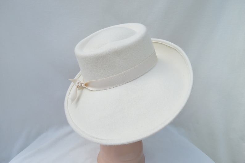 May include: A cream-colored hat with a wide brim and a cylindrical crown. A cream-colored ribbon is wrapped around the base of the crown and tied in a bow on the side. The hat is made of a soft, felt-like material.