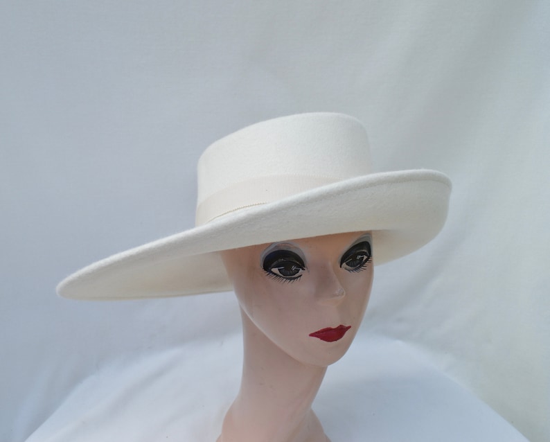 May include: A cream-colored hat with a wide brim and a flat crown. The hat features a light beige band around the base of the crown. The hat is displayed on a mannequin head with painted features, including dark eyes and red lips.