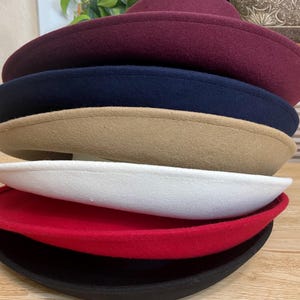 May include: A stack of felt hats in various colors, including burgundy, navy, tan, white, red, and black. The hats have a classic, rounded shape with a slightly upturned brim. The hats are stacked on top of each other.