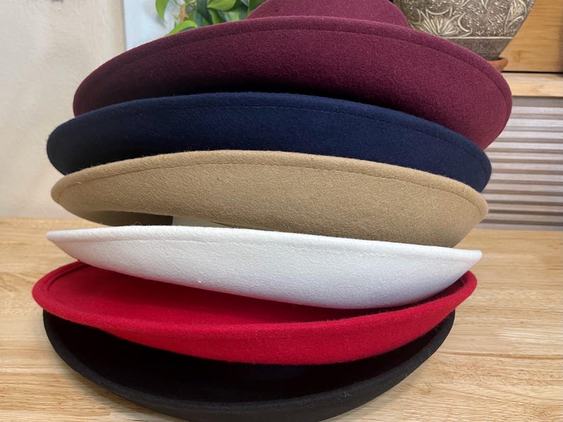 May include: A stack of felt hats in various colors, including burgundy, navy, tan, white, red, and black. The hats have a classic, rounded shape with a slightly upturned brim. The hats are stacked on top of each other.