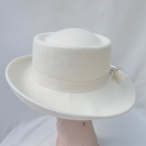 May include: A cream-colored hat with a wide brim and a cylindrical crown. A cream-colored ribbon is wrapped around the base of the crown, and a small bow is attached on the side. The hat is made of a soft, felt-like material.