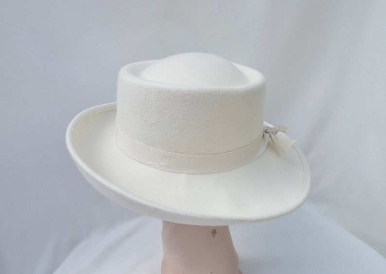 May include: A cream-colored hat with a wide brim and a cylindrical crown. A cream-colored ribbon is wrapped around the base of the crown, and a small bow is attached on the side. The hat is made of a soft, felt-like material.