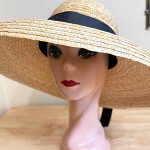 May include: A wide-brimmed straw hat with a black ribbon band. The hat is displayed on a mannequin head with red lipstick. The hat's brim is approximately 18 inches in diameter, offering ample sun protection. The hat is a light beige color.