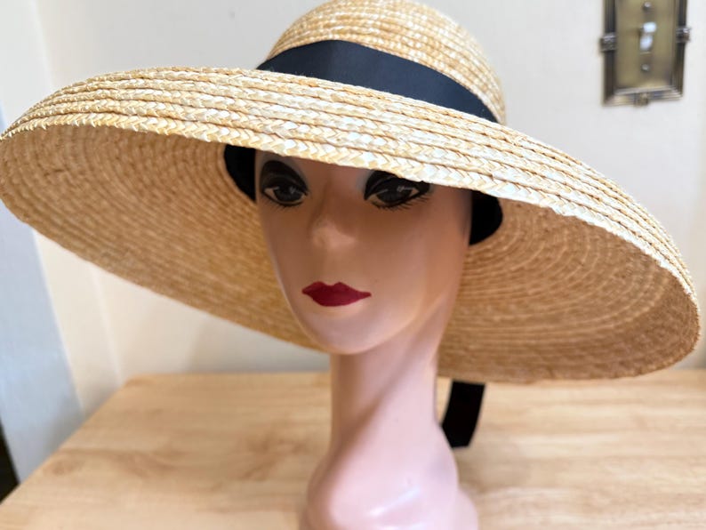 May include: A wide-brimmed straw hat with a black ribbon band. The hat is displayed on a mannequin head with red lipstick. The hat's brim is approximately 18 inches in diameter, offering ample sun protection. The hat is a light beige color.