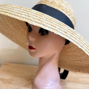 May include: A wide-brimmed straw hat with a black ribbon band. The hat is made of woven straw in a natural beige color. The hat is displayed on a mannequin head. The hat's brim is wide, providing ample shade.