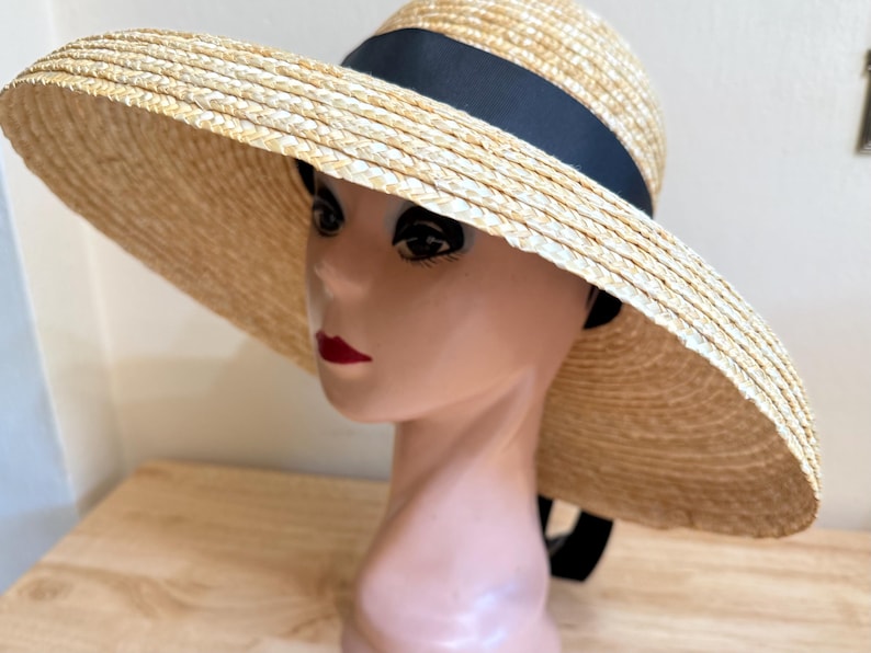 May include: A wide-brimmed straw hat with a black ribbon band. The hat is made of woven straw in a natural beige color. The hat is displayed on a mannequin head. The hat's brim is wide, providing ample shade.