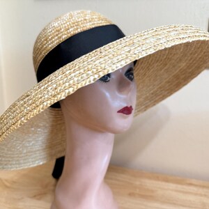 May include: A wide-brimmed straw hat with a black ribbon band. The hat is a natural beige color and has a large brim. The hat is on a mannequin head with red lipstick.