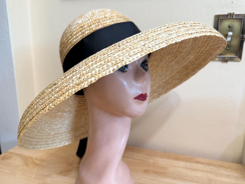 May include: A wide-brimmed straw hat with a black ribbon band. The hat is a natural beige color and has a large brim. The hat is on a mannequin head with red lipstick.