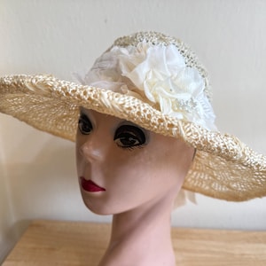 Puede incluir: Un sombrero para el sol color crema con ala ancha. El sombrero está hecho de paja tejida y presenta un arreglo decorativo de flores y adornos de tela blanca en la corona. El sombrero se exhibe en una cabeza de maniquí.