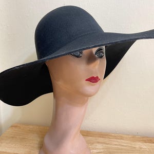 Könnte beinhalten: Ein schwarzer Filzhut mit breiter Krempe und leicht abgerundeter Krone. Der Hut wird auf einem Mannequin-Kopf getragen.