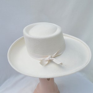 May include: A white hat with a wide brim and a cylindrical crown. A cream-colored ribbon is tied around the base of the crown and tied into a bow. The hat is made of a soft, felt-like material.