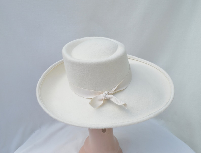 May include: A white hat with a wide brim and a cylindrical crown. A cream-colored ribbon is tied around the base of the crown and tied into a bow. The hat is made of a soft, felt-like material.
