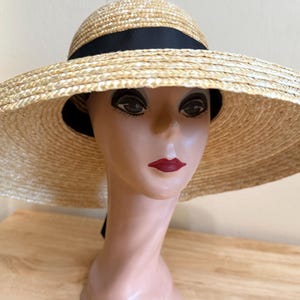 May include: A wide-brimmed straw hat with a black ribbon band. The hat is displayed on a mannequin head with painted features, including dark eyes and red lips. The hat's brim is wide, providing ample shade.