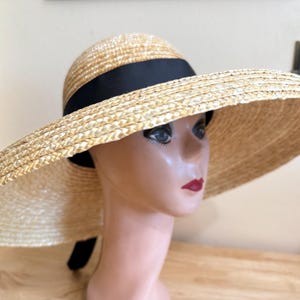 May include: A wide-brimmed straw hat with a black ribbon band. The hat has a rounded crown and a large, floppy brim. The hat is displayed on a mannequin head.