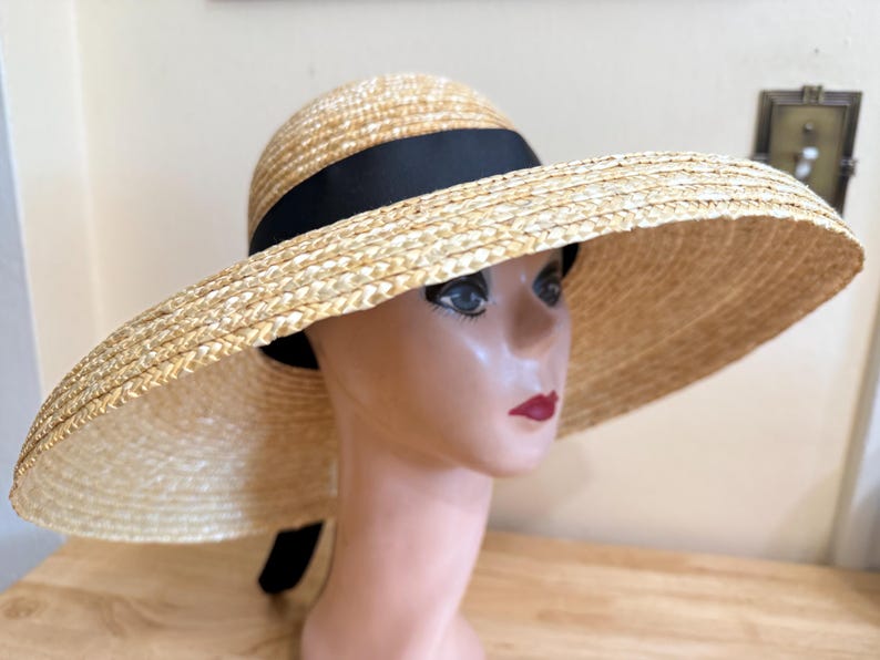 May include: A wide-brimmed straw hat with a black ribbon band. The hat has a rounded crown and a large, floppy brim. The hat is displayed on a mannequin head.