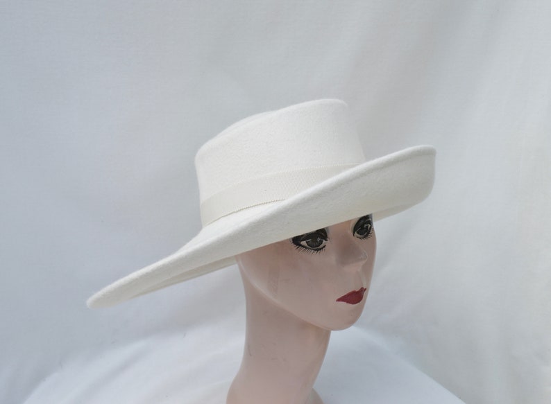 May include: A white fedora-style hat with a wide brim and a matching ribbon band. The hat is displayed on a mannequin head with red lipstick. The hat's brim is approximately 4 inches wide.