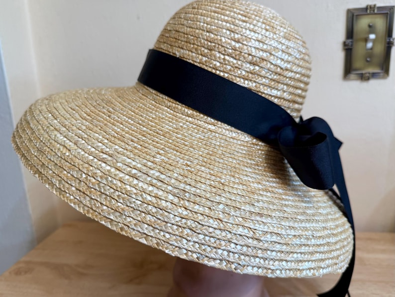 May include: A straw sun hat with a wide brim and a black ribbon tied in a bow. The hat is made of woven straw and has a classic, elegant design. The black ribbon adds a touch of sophistication to the hat.