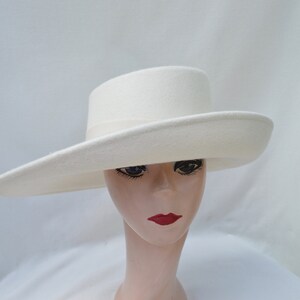 May include: A cream-colored, wide-brimmed hat with a flat crown and a ribbon band. The hat is made of a soft, felt-like material. The brim measures approximately 4 inches wide. The hat is displayed on a mannequin head with red lipstick.