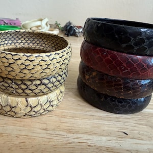 May include: Two stacks of circular bangle bracelets. The left stack consists of three light beige bangles with a dark, scale-like textured pattern. The right stack features four bangles in varying shades of dark red, brown, and black with a smooth finish.