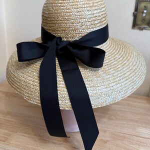 May include: A straw hat with a wide brim and a black ribbon tied in a bow. The hat has a rounded crown and a dark ribbon encircling the base of the crown. The ribbon is tied into a large bow on the front, with long streamers.