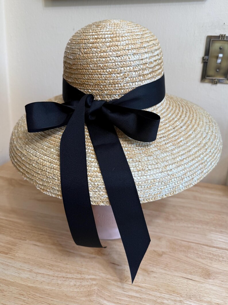 May include: A straw hat with a wide brim and a black ribbon tied in a bow. The hat has a rounded crown and a dark ribbon encircling the base of the crown. The ribbon is tied into a large bow on the front, with long streamers.