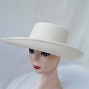 May include: A cream-colored hat with a flat brim and a cylindrical crown. The hat is made of a soft, felt-like material. The hat is displayed on a mannequin head.