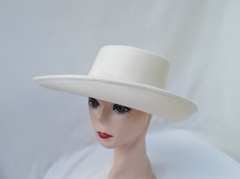 May include: A cream-colored hat with a flat brim and a cylindrical crown. The hat is made of a soft, felt-like material. The hat is displayed on a mannequin head.