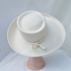 May include: A cream-colored hat with a wide brim and a cylindrical crown. A cream-colored ribbon is tied around the base of the crown in a bow. The hat is displayed on a pink hat stand.