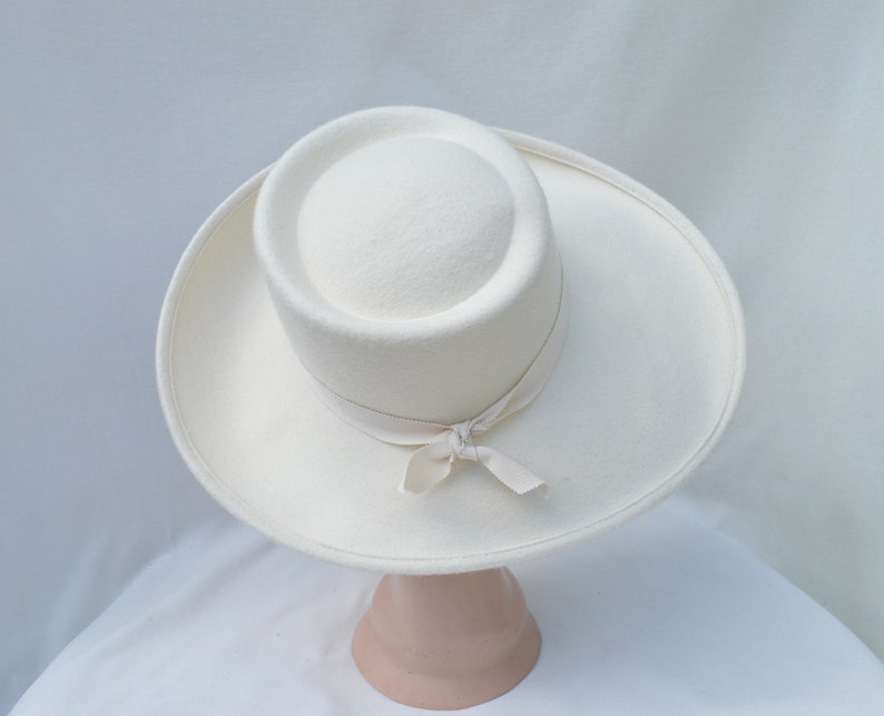 May include: A cream-colored hat with a wide brim and a cylindrical crown. A cream-colored ribbon is tied around the base of the crown in a bow. The hat is displayed on a pink hat stand.