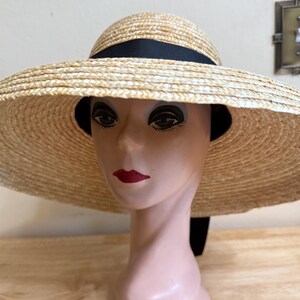 May include: A wide-brimmed straw hat with a black ribbon band. The hat is displayed on a mannequin head with red lipstick. The hat's brim is approximately 18 inches in diameter, offering ample sun protection. The hat is a classic accessory.