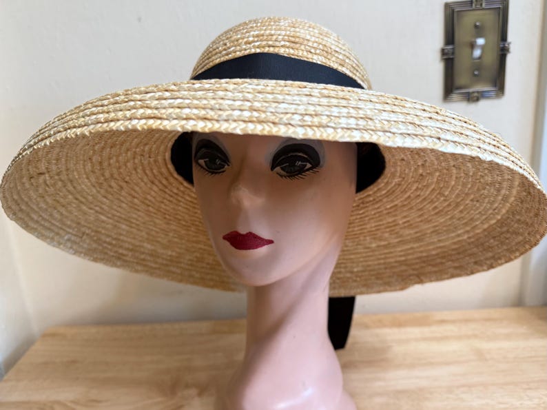 May include: A wide-brimmed straw hat with a black ribbon band. The hat is displayed on a mannequin head with red lipstick. The hat's brim is approximately 18 inches in diameter, offering ample sun protection. The hat is a classic accessory.