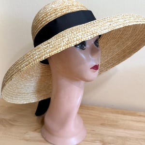 May include: A wide-brimmed straw hat with a black ribbon band. The hat is displayed on a mannequin head. The hat's brim is wide, and the straw has a natural, light tan color. The black ribbon adds a contrasting accent.