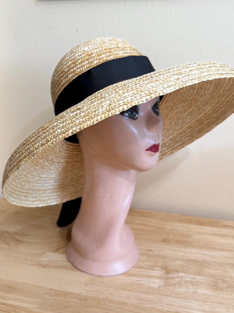 May include: A wide-brimmed straw hat with a black ribbon band. The hat is displayed on a mannequin head. The hat's brim is wide, and the straw has a natural, light tan color. The black ribbon adds a contrasting accent.