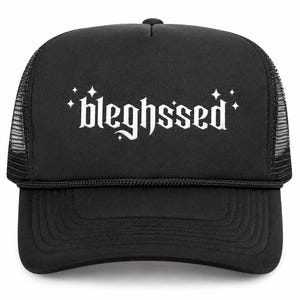May include: A black trucker hat featuring a foam front panel and mesh back. The front displays the white text "bleghssed" in a gothic-style font, accented by small four-pointed star icons positioned above and below the lettering.