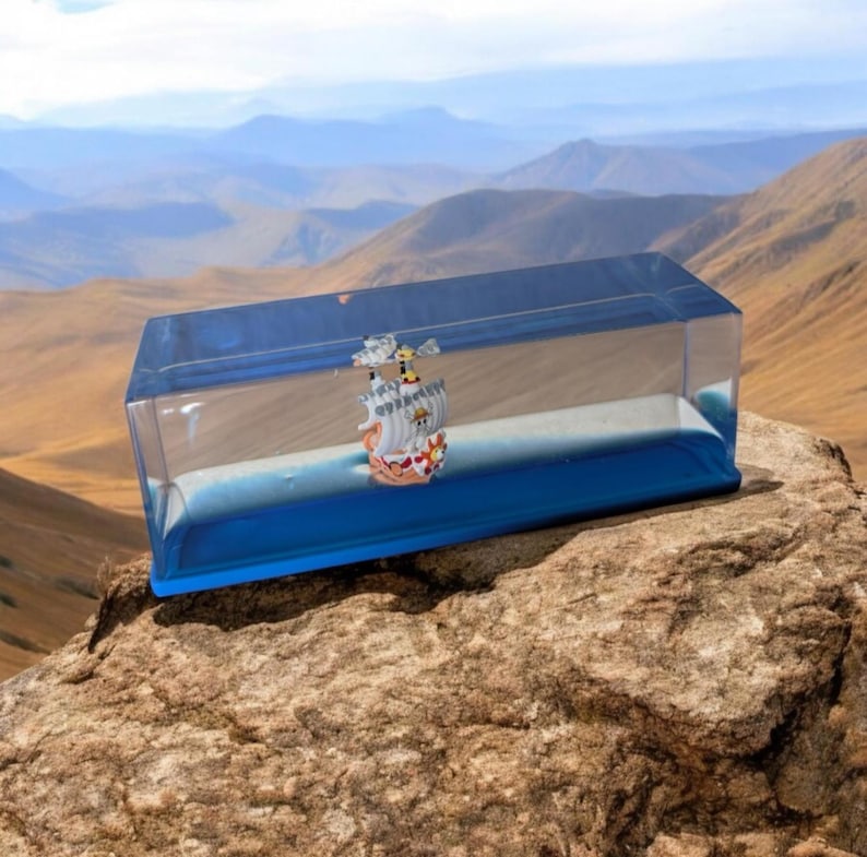 May include: A clear, rectangular hourglass with a blue base and a miniature ship inside. The ship is detailed with colorful sails and flags. The hourglass is filled with blue liquid and white sand, set against a mountain backdrop.