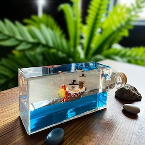 May include: A rectangular clear glass bottle filled with blue liquid and a miniature ship figurine inside. The ship features a lion-shaped bow, a white sail with a skull emblem, and a brown mast. The bottle is sealed with a cork stopper.