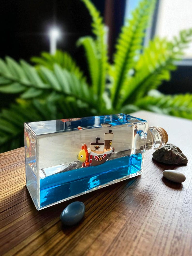 May include: A rectangular clear glass bottle filled with blue liquid and a miniature ship figurine inside. The ship features a lion-shaped bow, a white sail with a skull emblem, and a brown mast. The bottle is sealed with a cork stopper.