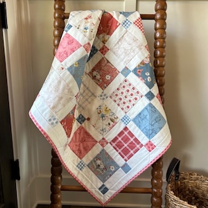 May include: A patchwork quilt with a variety of floral and plaid patterns in shades of pink, blue, and red. The quilt is draped over a wooden ladder. The quilt's dimensions are not visible.