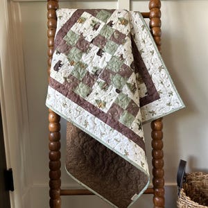 May include: A handmade quilt with a patchwork design featuring bears, trees, and floral patterns in shades of brown, green, and white. The quilt is draped over a wooden ladder, showcasing its intricate details and craftsmanship. The quilt's dimensions are not visible.