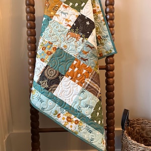May include: A patchwork quilt with a variety of patterns and colors, including horses, floral designs, and geometric shapes. The quilt is draped over a wooden ladder. The color palette includes teal, orange, brown, and white.