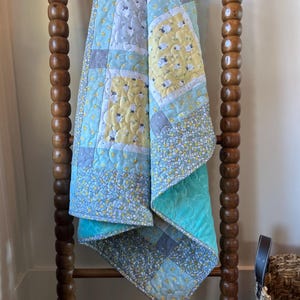 May include: A handmade quilt with a patchwork design, featuring squares of light blue, yellow, and gray fabrics. The quilt is adorned with sheep and floral patterns. It is draped over a wooden ladder, showcasing its intricate stitching and color palette.