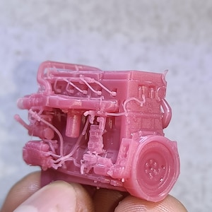 Könnte beinhalten: Ein kleines, detailliertes rosa Motormodell mit verschiedenen Komponenten wie Rohren und einem Schwungrad. Das Modell wird in einer Hand gehalten, um seine Größe zu zeigen. Der Motor hat eine einheitliche rosa Farbe und zeigt sein komplexes Design.