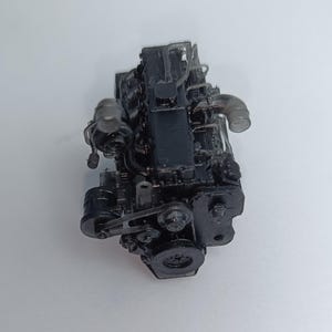 May include: A detailed, black model engine with various components, including pipes, belts, and a flywheel. The engine is set against a plain white background, showcasing its intricate design and mechanical features.