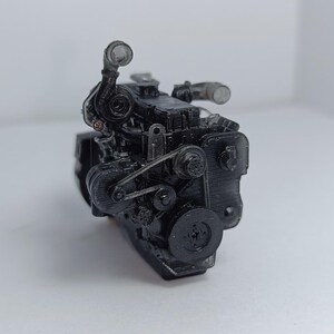 May include: A black engine model with intricate details, including belts, pulleys, and tubes. The engine is set against a plain white background, showcasing its mechanical components. The model appears to be a miniature replica of an internal combustion engine.