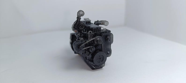 May include: A black engine model with intricate details, including belts, pulleys, and tubes. The engine is set against a plain white background, showcasing its mechanical components. The model appears to be a miniature replica of an internal combustion engine.