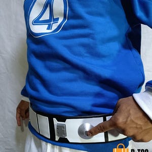 Puede incluir: Una sudadera azul con un logotipo circular blanco con el número 4. Un cinturón blanco y negro con un botón plateado está siendo presionado por un dedo. El cinturón se usa sobre pantalones blancos.
