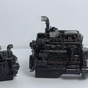 Puede incluir: Dos modelos de motores negros se muestran sobre un fondo blanco. El motor más grande tiene detalles intrincados, incluyendo tuberías y varios componentes. El motor más pequeño refleja el diseño, mostrando el mismo nivel de detalle.