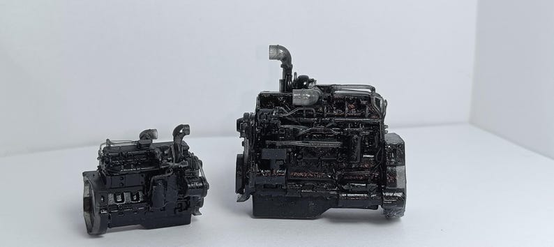 May include: Two black model engines are displayed against a white background. The larger engine has intricate details, including pipes and various components. The smaller engine mirrors the design, showcasing the same level of detail.