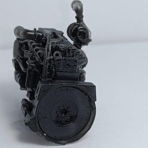 May include: A detailed, black model of an engine, showcasing intricate mechanical components. The model features a cylindrical flywheel and various pipes and tubes, all rendered in a matte black finish. The engine is positioned against a plain white background.